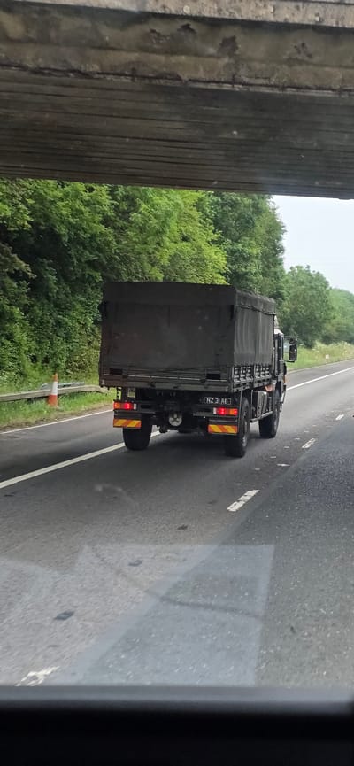 A dark-colored, canvas-covered military-style truck is driving on a highway. The truck has a license plate that reads "NZ 31 AB". The truck is on the right side of the road, with trees and foliage on the left and right sides of the highway. A concrete overpass is visible at the top of the image, casting a shadow over the road. The weather appears to be overcast. The location is identified as Barton, United Kingdom.