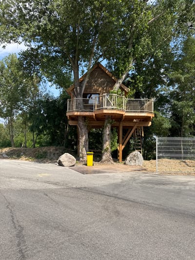 The image depicts a treehouse nestled within the branches of two large trees. The treehouse has a wooden structure with a balcony and a pitched roof. A ladder is visible, providing access to the treehouse. In front of the treehouse, there is a large rock and a yellow trash can. A metal fence runs along the right side of the image. The scene is set outdoors, with lush green trees and foliage surrounding the treehouse, under a clear blue sky. The location is identified as Grenay, France.