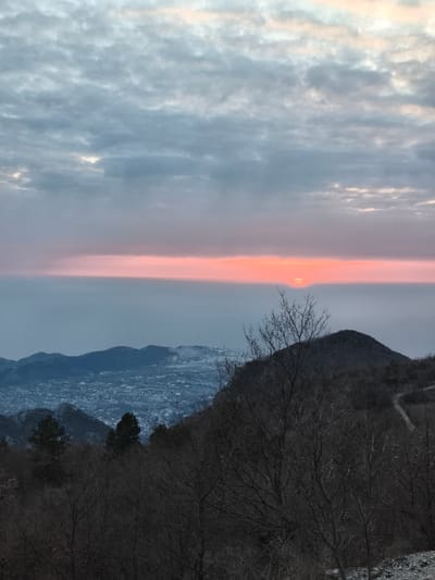 A panoramic landscape photograph captures a twilight scene from Mali Mikulići, Montenegro. The upper two-thirds of the image display a sky dominated by grey stratocumulus clouds, transitioning to a distinct horizontal band of vibrant orange-red light at the horizon. The sun's upper arc is partially visible within this band, subtly obscured by atmospheric haze or distant clouds. Below the horizon light, a wide, hazy expanse of light blue-grey, consistent with the Adriatic Sea, stretches across the mid-distance. Further into the midground, a sprawling urban area is visible, characterized by numerous structures and diffused artificial lighting, situated at the base of dark, undulating mountain ranges. The foreground and right midground are occupied by a steep, rocky hillside, sparsely covered with dormant deciduous trees and shrubs, their bare branches silhouetted against the lighter background. A narrow, unpaved track is discernible winding upwards along the far right slope of this hillside. No human figures, animals, or explicit actions are present within the frame.