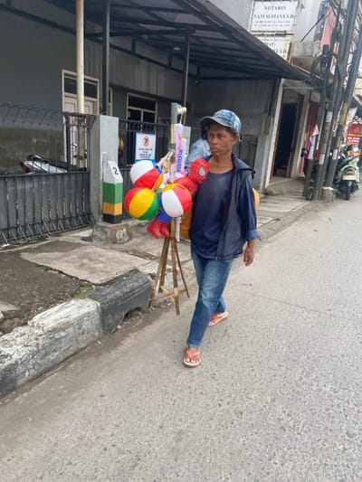 A man is walking down a paved street in Bojongsoang, Indonesia (latitude: -6.972, longitude: 107.636). He is carrying a stand with inflatable toys, including a Spiderman, a colorful ball, and a striped ball. He is wearing a blue shirt, blue jeans, a blue hat, and orange sandals. He is walking on a sidewalk next to a concrete curb and a black iron fence. On the opposite side of the street, there are buildings with a sign for a notary's office. There is a motorcycle parked on the side of the street.