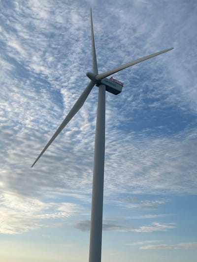 A low-angle, full shot captures a towering wind turbine against a vast, cloudy sky. The turbine, a Siemens model, stands tall with three long, white blades extending outwards. The nacelle, the housing at the top of the turbine, is visible, featuring the Siemens logo and a small red railing around an observation deck. The sky is filled with wispy, white clouds scattered across a bright blue expanse, creating a dynamic backdrop. The horizon, a pale line of light, suggests either the sea or a distant landscape. The scene appears to be located in Nasva alevik, Estonia, a coastal area known for its wind energy infrastructure. There are no people visible in the image.