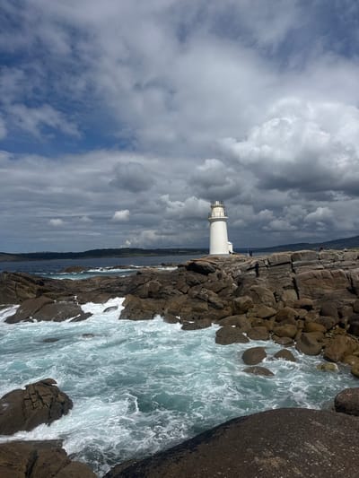 The image shows a lighthouse on a rocky coast in Muxía, Spain. The lighthouse is white and tall, with a black top. It is located on a small island, surrounded by crashing waves. There are a few people standing on the rocks near the lighthouse. The sky is a cloudy blue, with large, fluffy white clouds. The water is a bright turquoise color, and the waves are white and foamy. The rocks are brown and grey. The image appears to have been taken on a sunny day with some cloud cover. The image was taken from a point of view looking directly at the lighthouse and the rocky coast. The photograph is well-composed, and the colors are vibrant. The overall tone of the image is peaceful and serene. The lighthouse is located on the coast of the Atlantic Ocean, at the coordinates (43.113, -9.221) which is in the town of Muxía in Spain.