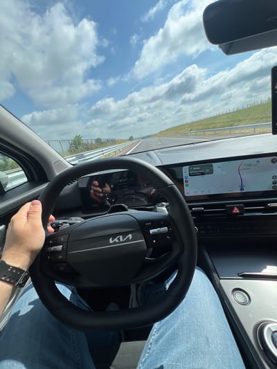The image captures the interior of a modern car, likely a Kia due to the prominent logo on the steering wheel, as it travels on a highway under a bright, partly cloudy sky. The perspective is from the driver's seat, showing the driver's right hand holding the steering wheel and their light blue jeans.

**Subjects and Objects:** The main subjects are the car's interior components and the road ahead. Key objects include the black leather-wrapped steering wheel with the "Kia" logo and control buttons (one labeled "MODE"), a widescreen digital dashboard and infotainment system, and a "START STOP" engine button on the right side of the dashboard. Outside the car, a multi-lane road, guard rails, grassy verges, and a distant tree line are visible under a sky dotted with white clouds. A wristwatch is visible on the driver's wrist.

**Activity:** The scene depicts the activity of driving a car, evidenced by the driver's engaged posture, the active navigation system, and the moving road environment.

**Notable Details:** The time of day is daylight, with clear visibility. The weather is partly cloudy. The infotainment screen displays navigation information, showing a route with "94 km" remaining to a destination, and listing "Варна / Varna" and "Слънчев бряг" (Sunny Beach) as key locations. The screen also indicates a current time of "13:29", an estimated remaining travel time of "2:59", a total remaining distance of "230" km, and an estimated arrival time of "16:33". The digital instrument cluster, partially visible, shows readings such as "72.1", "0:42", "86", "2.4 x 1000 rpm" (2400 RPM), and "D7" (indicating the car is in 7th gear, likely automatic). Several speed-related numbers are also visible in the cluster or its reflection, including "133", "140", and "1356". A red hazard light button is visible on the center console. The visible locations confirm the setting is in Bulgaria, in the region of Chokoba. No specific emotions are discernible.