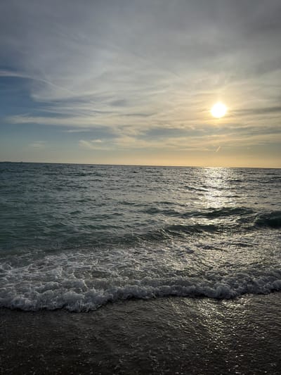 A serene seascape captures a body of water under a sky adorned with scattered clouds and a bright, low-hanging sun. The sun, a brilliant orb, casts a shimmering reflection on the rippling surface of the water, creating a path of light that stretches towards the viewer. The water itself is a deep, inviting blue-green, with gentle waves breaking along the shore, leaving a foamy white surf. The foreground is a dark, wet sand beach, glistening with moisture from the receding tide. In the distance, the horizon is a sharp, clean line separating the water from the sky. A faint contrail from an aircraft streaks across the upper left quadrant of the sky, adding a touch of human presence to the otherwise natural landscape. The scene evokes a sense of tranquility and the vastness of nature.