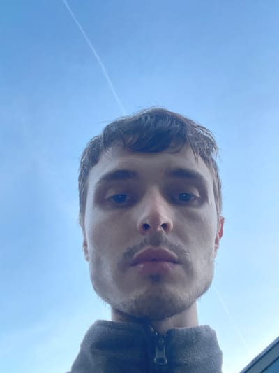A low-angle, close-up selfie shows the face of a person against a bright blue sky with a faint contrail. The person has short, dark, messy hair and a light mustache and stubble. Their eyes are dark, and their lips are closed. They are wearing a dark gray, fuzzy jacket with a zipper that is partially visible at the bottom of the frame. The background is a clear blue sky with a single, thin white contrail stretching diagonally from the upper left to the lower right. The image appears to be taken outdoors in Genk, Belgium.