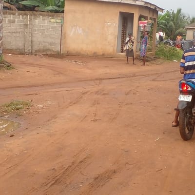 The image captures a reddish-brown dirt road intersection in Ijotan, Nigeria, under an overcast sky. In the foreground on the right, a person wearing a blue and white striped shirt and brown sandals is seated on a blue and black motorcycle, viewed from the rear; its license plate reads "OSUN M-7170J". The dirt road is extensively marked with tire tracks and some footprints, and contains small puddles of water. To the right of the intersection, two women stand near a single-story, light brown building with a dark recessed doorway. One woman wears a light-colored top and a dark skirt, holding a bag. The second woman, in a patterned dress, carries a grey plastic basket filled with items balanced on her head. To the left, a grey concrete block wall displays a sign partially legible as "IJUE - STIL", beyond which lush green foliage, including banana plants, is visible. Several indistinct figures are present further in the background to the right.