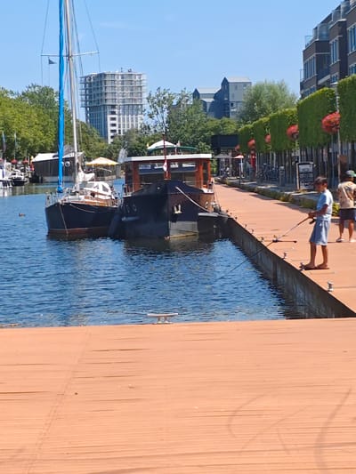 This image depicts a canal-side scene in the city of Tilburg, Netherlands, likely in the Spoorzone area given the modern architecture and industrial heritage evident. In the foreground, a wooden pier extends towards the viewer, with several cleats visible along its edge. To the left, a dark-hulled sailboat with a white cabin and a tall mast is moored in the blue water. Adjacent to it, a larger, dark-hulled boat, possibly a houseboat, is also docked.

On the pier, a young boy, dressed in a light blue t-shirt and grey shorts, is actively fishing with a rod. He stands with his back to the viewer, facing the water. Further down the pier, another person, wearing a patterned shirt and shorts, is also visible, seemingly observing or engaged in a similar activity. In the background, a row of modern apartment buildings with balconies lines the opposite side of the canal, interspersed with trees and some green hedges with red flowers. The sky is clear and blue. Several other boats are visible further down the canal, suggesting a bustling waterway. The overall atmosphere is one of a pleasant summer day, with recreational activities taking place along the water.
