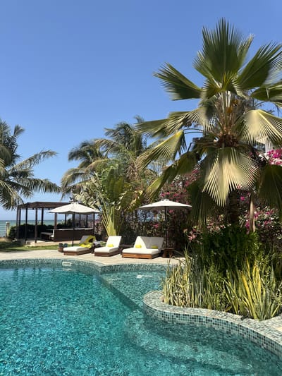 The scene is an outdoor leisure area in Ngaparou, Senegal, beside a swimming pool with turquoise water. The pool edge is decorated with blue-green mosaic tiles. There are lounge chairs with white cushions and yellow towels, shaded by white umbrellas. Tropical vegetation including palm trees and flowering bushes surrounds the area. In the background, a pergola structure overlooks the ocean. The sky is clear blue.