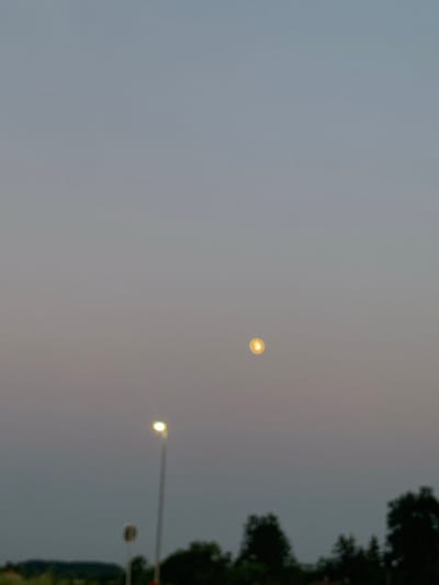 The image captures a twilight sky over a landscape in Ub Municipality, Serbia. A single street light is visible on the left, illuminating a portion of the scene. In the center of the frame, a bright, circular object, possibly a street light or a distant lamp, is suspended in the air. The horizon line is marked by silhouetted trees and foliage, suggesting a rural or semi-urban setting. The sky transitions from a pale blue at the top to a warmer, hazy tone near the horizon.