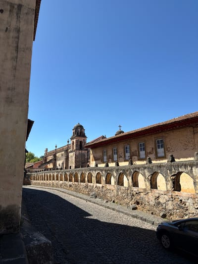 The image displays an outdoor scene dominated by historical architecture under a clear blue sky. The photo appears to be taken with a vertical orientation (portrait mode) but rotated 90 degrees counter-clockwise, making the sky appear on the left and the ground on the right.
In the mid-ground and extending into the background, a long, multi-story building with a textured, light-colored facade (possibly stucco or aged stone) stretches horizontally across the frame. The upper part of this building features multiple small, rectangular windows with white frames. Below these windows, a series of arched openings or alcoves form a lower level or a retaining wall-like structure, also made of rough-hewn stone or aged material. The roofline of this building, visible at the top, has a reddish-brown tiled appearance. A small cross can be seen on one of the roof peaks towards the center of the image.
Further in the background, behind the main building and slightly to the left, a more ornate structure with a domed tower or steeple is visible, suggesting a church or a similar grand edifice. This structure also appears to be made of stone and shows signs of age.
In the foreground, on the right side of the image (which would be the bottom if correctly oriented), a dark-colored, possibly paved or cobbled street or pathway is present. A significant portion of this ground is covered in deep, elongated shadows, indicating a bright sunny day with the sun positioned to the left of the scene. Part of a dark-colored car, specifically its front bumper, wheel, and side, is visible parked on the street in the upper right corner of the frame.
The sky, occupying the entire left half of the image, is a vibrant, cloudless blue. In the immediate foreground, at the very bottom left, a low, light-colored concrete or stone wall is partially visible, suggesting the viewpoint is slightly elevated or looking over another structure.
The overall impression is one of a historic town or city, likely in a sunny climate, characterized by aged stone buildings and traditional architecture. No text is visible in the image.