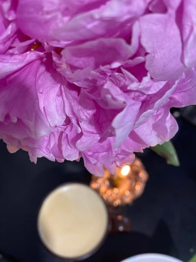 An extreme close-up shot focuses on the delicate, layered pink petals of a peony flower, with hints of yellow stamens visible within its folds. Below the flower, a blurred background reveals a warm, glowing candle surrounded by bokeh lights, casting an ethereal ambiance. To the left, the top portion of a light-colored beverage in a glass is visible, also out of focus. The scene suggests a serene and intimate moment. The image does not contain any identifiable landmarks or details that would place it in Podgorodnyaya Pokrovka, Russia.