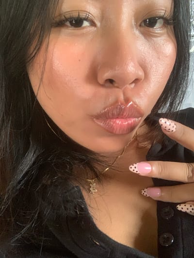 A close-up, portrait-style photograph shows a person's face and upper body, with a particular focus on their lips and hands. The person is wearing dark clothing and has long, dark hair. Their lips are coated in a glossy product, appearing plump and shiny. The person's right hand is visible, displaying manicured nails decorated with a pink base color and black polka dots. A delicate gold necklace with a charm is also visible around the person's neck. The background is out of focus, with some hints of reflective surfaces, suggesting an indoor setting. The lighting is soft and appears to highlight the dewy texture of the person's skin and lips. The image is framed tightly, cropped to emphasize facial features and cosmetic details. There is no indication of the city of Jajag, Indonesia in the image.