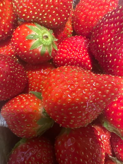 The image is a close-up, top-down view of a pile of fresh, ripe strawberries. The strawberries are red and plump, with numerous small, yellow seeds dotting their surfaces. Some of the strawberries still have their green leafy tops attached. The lighting suggests it is daytime, and the setting appears to be indoors, possibly a kitchen or a market stall, given the focus on the fruit. There is no visible text in the image. The scene evokes a sense of freshness and natural abundance.