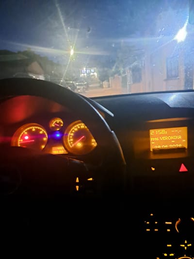 The image shows the interior of a car at night. The dashboard is illuminated with the driver's perspective. The main focus is on the instrument cluster and the car's radio display. The instrument cluster features two prominent circular gauges, likely the speedometer and tachometer, with their needles indicating specific values. A smaller, circular gauge is positioned above them. A digital display below the gauges shows "400.7" and "466 165".
To the right of the instrument cluster, the car's radio display is illuminated, showing "21:58" in the top left corner. Below this, it reads "FM6 VERONIKA". Underneath this, it displays "RDS" and "28.06.2026". There are several buttons around the radio display and further down on the dashboard, some of which are also illuminated. A red warning triangle is visible on the right side of the dashboard.
The view through the windshield shows a street scene at night, with streetlights and illuminated houses visible. Cars are parked or driving in the background. The windshield has some smudges and streaks. The overall lighting suggests it is dark outside, and the interior lights of the car are on. The location is identified as Ignatievo, Bulgaria.