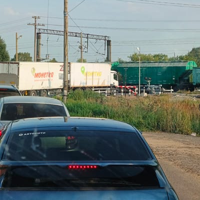 In Kambarka, Russia, a train crossing is depicted with a line of cars stopped. The foreground shows the rear of a dark blue car, featuring a sticker that reads "Автосити" (Avtositi) on its tinted rear window and a row of red brake lights. To the left and slightly behind it, another car's dark rear window is visible.

In the mid-ground, a white truck with the logo and text "МОНЕТКА" (Monetka) is stopped, implying it is a delivery vehicle for the "Monetka" grocery store chain. Next to the white truck is a long, green train carriage, with some visible Cyrillic text and numbers, including "049 07817." A red and white striped railway crossing barrier is lowered, indicating an approaching or passing train.

The background features power lines and poles, typical of a railway infrastructure. The environment is characterized by tall green grass and other vegetation on the sides of the dirt and gravel road leading to the crossing. The sky is clear and blue, suggesting daylight. The overall scene captures a moment of temporary halt due to the train crossing.
