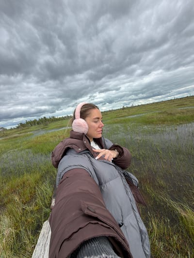 A young woman is lying on a wooden plank, her eyes closed and a peaceful expression on her face. She is wearing a brown and gray puffer jacket, pink earmuffs, and a grey knitted sweater. Her nails are painted white. She is outdoors in a marshy landscape. The ground is covered with green grass and patches of water, suggesting a wetland environment. In the background, there is a line of trees on the horizon, and a vast expanse of cloudy sky fills the upper portion of the image. The clouds are gray and streaky, indicating overcast weather. The overall mood is serene and tranquil, with the woman appearing to be enjoying a moment of rest in nature. The time of day is indeterminate due to the overcast conditions, but the lighting suggests it is daytime. The location context provided is Мёрскі сельскі Савет, Belarus, which aligns with a marshy, temperate climate with potential for dense cloud cover. No visible text is present in the image.