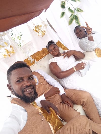 The image displays three individuals, two females and one male, seated on an ornate white and gold couch positioned under a decorated canopy in Ugwolawo, Nigeria. The male, positioned in the foreground on the left, has a beard and short dark hair, wearing a white long-sleeved shirt, a tan waistcoat with gold buttons, and matching trousers. A gold chain is visible under his waistcoat, and a gold ring is on his left ring finger. He smiles directly towards the camera. The central female, behind the male, wears a white off-the-shoulder lace wedding gown, a gold necklace, a gold bracelet on her right wrist, and a gold ring on her left ring finger. She holds a light blue, patterned clutch bag in her lap and forms a peace sign with her right hand while smiling at the camera. The second female, located in the upper right background, wears a long-sleeved white garment with embroidered details and multiple bangles on her left wrist. She also forms a peace sign with her left hand and smiles. The background features white draped fabric adorned with artificial green leaves and gold-colored decorative elements. A dark brown fabric canopy covers the upper left portion of the scene.