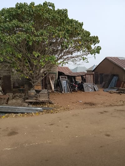 The image displays an outdoor scene in Minting Chip, Nigeria, under an overcast sky. A brown, unpaved road or compacted earth path occupies the foreground. To the left, a large, deciduous tree with dense green and yellowish foliage stands over reddish-brown dirt, which is scattered with dry leaves.

Behind the tree, a structure made of grey concrete blocks is partially visible. Adjacent to it, centrally positioned, is a lean-to workshop or storage area with a corrugated metal roof. This area contains multiple stacked metal doors, door frames, and metal sheets, some featuring embossed patterns. A red and black motorcycle is parked beneath the lean-to. To the far right, a building with mud-colored walls and a rusted corrugated metal roof has additional metal sheets and lumber leaning against its exterior. Distant structures with similar roofing are visible in the background.

On the ground in the lower left, stacks of long, rectangular metal profiles are present. Under the large tree, a wooden structure resembling a bench or platform supports a piece of equipment with a funnel-shaped top, possibly a rudimentary grinder. No individuals are present, and no actions or interactions are depicted.