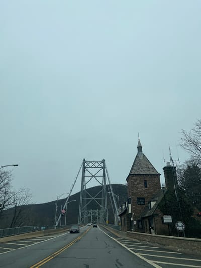 The image captures an overcast daytime scene on the eastern approach of the Bear Mountain Bridge in the Town of Cortlandt, United States of America. A two-lane asphalt road, marked by double yellow center lines, leads westward onto the steel suspension bridge. Three passenger vehicles are visible on the roadway, proceeding away from the viewer towards the bridge's main span. To the right of the road stands a multi-story stone building with a prominent grey conical spired roof and multiple windows. The building's right facade and an adjacent chimney are partially covered in green ivy, with an antenna mast mounted near the chimney. A circular white sign on the building's side partially reads "NEW YORK STATE" and "BEAR MOUNTAIN." Bare deciduous trees flank both sides of the road, and tree-covered hills rise in the background beneath a uniformly grey, cloud-laden sky. An American flag is visible on a pole near the bridge structure on the left.