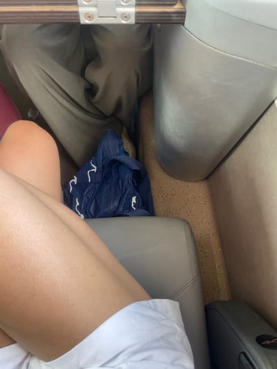 The image depicts the interior of a public transport vehicle, likely a train, located in or en route to/from Afragola, Italy. The view is from a seated, first-person perspective, looking forward and downwards.

In the immediate foreground, the bare lower thighs and knees of an individual, possibly female, are visible, wearing white shorts or a skirt. The skin tone is fair, with visible hair follicles. A light-grey upholstered seat armrest is partially visible on the lower right.

Directly ahead, the lower torso and legs of a second individual are visible, seated opposite or in an adjacent row. This person is wearing dark, possibly olive-green or grey, trousers. Below their legs, light-colored, possibly beige or white, patterned footwear is discernible on the floor.

Between the two individuals, on the floor, a crumpled dark blue bag with white, irregularly shaped patterns or logos is present. The floor surface is light brown or beige with dark speckles, indicative of a durable, textured material.

Above the second individual's legs, a wooden or wood-laminate surface with a silver-colored metal hinge mechanism is visible, consistent with a deployed tray table. To the right, a large, curved, matte grey structural element of the vehicle's interior is present, likely the base of a seat or a wall panel, displaying minor scuff marks. The overall environment is functional and utilitarian, typical of a train cabin.