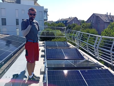 A man in red shorts and a grey t-shirt stands on a rooftop in Murten, Switzerland, facing a row of solar panels. He is wearing sunglasses and gloves, and appears to be holding a cigarette. The rooftop is lined with a silver railing, and in the background, residential buildings and lush green trees are visible. The sky is clear and blue.