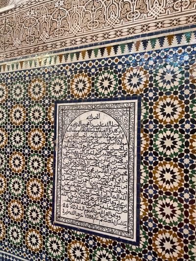 This image depicts a section of an intricately decorated interior wall in Moulay Idriss Zerhoun, Morocco. The upper part of the wall features light-toned, carved stucco plasterwork with detailed arabesque and geometric relief patterns. Below this, a horizontal band of glazed tiles forms a frieze, incorporating dark blue, green, and white geometric motifs, including upward-pointing triangular elements and small circular accents. The majority of the wall surface is covered in traditional Moroccan zellij mosaic tiles, exhibiting complex and repetitive geometric patterns composed of stars, crosses, and other polygons in shades of white, dark blue, green, and brown. Embedded within this mosaic, slightly right of the vertical center, is a rectangular white plaque framed by a dark blue and white ornate border. This plaque contains multiple lines of black Arabic calligraphy, indicating a historical or dedicatory inscription. No individuals, actions, or interactions are present in the frame. The lighting suggests an indoor setting, illuminating the intricate details of the craftsmanship.