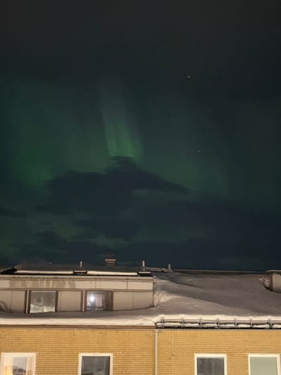 The image captures a nocturnal scene in Kiruna, Sweden, featuring a vibrant aurora borealis over urban rooftops. The upper half of the frame displays a dark night sky with prominent green light emissions characteristic of the aurora, appearing as several distinct vertical rays and diffused glows, particularly on the left and center-right. These auroral displays are partially obscured by dark, scattered clouds. Multiple small, bright celestial bodies are visible in the darker sky regions.
The lower half of the image presents the snow-covered roofs of at least two buildings. In the immediate foreground, the top section of a light-yellow brick building is visible, showing two distinct rectangular windows. The window on the lower left exhibits a faint internal artificial light source. Above and behind this structure, a flat-roofed building with a light-colored, possibly off-white, facade extends across the midground. This roof is thoroughly blanketed in snow, displaying uneven drifts and accumulations. Several dark, slender vertical structures, consistent with vents or small chimneys, protrude from the snow-covered surface. A single window on the left side of this light-colored building reflects a bright internal light source, while another window on the right remains dark. Further in the background, a segment of another snow-covered roof is discernible behind the flat-roofed building. No human subjects or active movements are visible.
