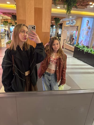 Two girls are taking a selfie in a mirror in a mall. One is holding the phone and wearing a black jacket. The other is standing behind her, wearing a brown jacket and jeans. The mall has multiple levels with stores and plants. The location is most likely Ol'ginskaya, Russia.