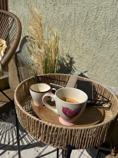 The image shows a patio scene, possibly in Mechkarevo, Bulgaria (location inferred but not directly visible). A round, woven, light brown table holds several items: two mugs, one with a pink base and a heart design and both appearing to contain coffee or a similar beverage, a mobile phone, a black geometric object (possibly a magazine rack), and a wooden tray. Pampas grass decorates the table's periphery, casting shadows on a textured, neutral-colored wall behind it. Part of a woven chair is visible in the lower left corner. Sunlight illuminates the scene, casting shadows and highlighting textures. A blanket rests partially visible on the chair. Part of a patterned rug is seen beneath the table.