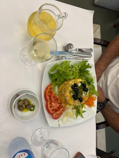 The scene depicts a restaurant table in Coruche, Portugal, covered with a white, textured tablecloth. A single individual's right arm is visible on the right side of the frame, wearing a white shirt and a black wristwatch.
Centrally placed on the table is a white rectangular plate containing a meal. The main component is a dish resembling shredded codfish with eggs and potatoes, garnished with whole black olives. This is surrounded by a fresh salad comprising green lettuce leaves, four slices of red tomato, shredded orange carrots, and thinly sliced white vegetables (likely onion or endive).
To the left of the main plate, two small white bowls are stacked; the top bowl holds green olives submerged in olive oil with visible herbs.
Beverages include a clear glass pitcher filled with a pale yellow liquid, presumably white wine, and a matching wine glass also containing the yellow liquid. Further down on the table, a plastic water bottle with a blue "LUS" branded label (likely Luso water) and an empty wine glass are visible.
A set of silver-colored cutlery, consisting of two forks, a knife, and a spoon, is neatly arranged on the tablecloth to the right of the filled wine glass. A dark brown object, possibly a phone or wallet, is partially visible near the empty wine glass.