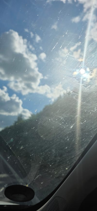 The image depicts a view through a car windshield, looking upwards at the sky. The windshield is visibly dirty and scratched, with numerous white speckles scattered across its surface. Bright sunlight is a prominent feature, creating lens flare and a strong glare that washes out parts of the image. The sky is a clear blue, dotted with fluffy white clouds. There is a suggestion of green foliage or trees in the reflection on the right side of the windshield, implying an outdoor setting. The upper right corner of the image shows a dark, textured surface which is likely part of the car's interior, possibly the dashboard or A-pillar, with a circular black object, possibly a speaker or sensor, also visible. The lighting indicates it is daytime, likely midday given the intensity of the sun. The scene evokes a sense of a casual drive on a sunny day, with the dirt and scratches on the windshield adding a touch of realism and perhaps a hint of neglect to the vehicle. No text is visible in the image.