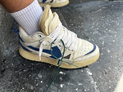 A white sneaker with blue accents is worn on a person's foot, which is covered by a white sock. The sneaker has a green plastic security tag attached to its laces. The shoe is resting on a speckled grey surface, possibly a step or pavement. The background is out of focus but appears to be an urban outdoor environment. The scene is photographed from a close-up perspective, focusing on the shoe. No specific location in Hamm, Germany can be determined from the image.