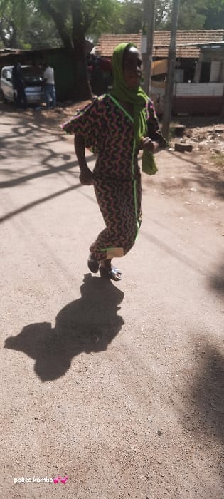 The image portrays a daytime outdoor scene located in Homa Bay, Kenya. The foreground features a dark-skinned individual, appearing female, in mid-stride on a dusty, unpaved road, moving from right to left. She is dressed in a long, patterned garment with a dark base and pink/orange geometric designs, accented with neon green trim along the V-neck and sides. A green headscarf covers her head, and she wears open-toed sandals. Her body casts a long, distinct shadow on the road surface to her left, indicating bright sunlight.
In the mid-ground to the left, a light-colored vehicle, consistent with a white van or car, is parked with its hood open. Two male individuals are positioned near the vehicle; one leans over the open engine compartment, and the other stands adjacent. Behind the vehicle, a large tree with green foliage is visible. To the right of the main subject, a utility pole extends upwards. The background reveals several simple structures with corrugated iron roofs, suggesting residential or small commercial buildings, interspersed with additional trees and dry terrain. The overall environment is characterized by dry earth and clear skies.