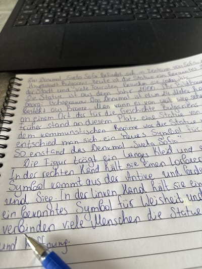 The image shows a page of a notebook with blue penmanship in German. The text describes a statue of "Sveta Sofia" located in the city center of Sofia, Bulgaria. The statue is made of bronze and is approximately 24 meters high. It was erected in 1900 and is situated on a spot where a statue of Georgi Chapkanov once stood. The statue represents a symbol of the communist regime and has come to represent Sveta Sofia. The figure is depicted wearing a long robe and holding a laurel wreath in its right hand, symbolizing victory. In its left hand, the figure holds a well-known symbol representing wisdom and hope. The provided context does not mention Groß-Gerau, Germany. The notebook page is lying on a dark surface, likely a laptop keyboard, partially visible at the top of the image. A blue pen is positioned in the lower left corner, obscuring some of the text.