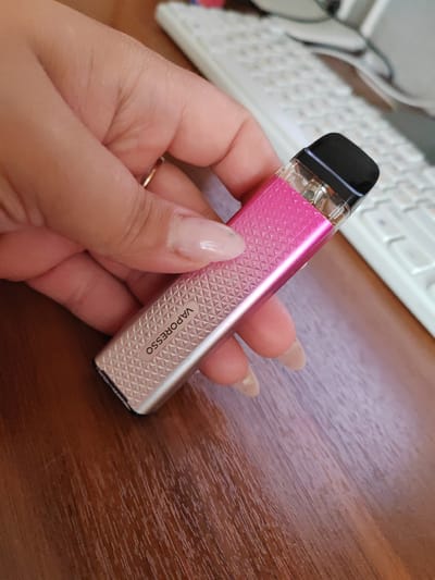 A close-up, eye-level shot shows a person's hand holding a pink and silver Vaporesso vape device. The device has a textured surface and the brand name is visible in black lettering. The hand has light-colored nail polish and a ring on one finger. The background includes a blurred white keyboard and a brown wooden surface, possibly a desk. The image is cropped to focus on the hand and vape device, with no discernible elements suggesting a location in Bol'shiye Klyuchishchi, Russia.