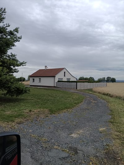 This media file depicts a rural scene in Dřevčice, Czechia. The primary subject is a white, single-story house with a red tile roof, featuring a chimney and several windows. A concrete fence encloses part of the property. The house is situated adjacent to a field of ripe wheat, which dominates the right side of the frame and extends to the horizon. To the left of the house, there is a patch of green grass and a large conifer tree. A gravel path curves from the foreground towards the house, suggesting it is a driveway.
The setting appears to be daytime, given the natural light, although the sky is overcast with heavy gray clouds, creating a muted and slightly somber atmosphere. The weather seems to be cloudy and potentially damp.
In the foreground, a portion of a vehicle's side mirror and a red element with visible white text are present, indicating the photo was taken from inside or next to a vehicle. The text on the red element is partially obscured and appears to be "TIZMAR" or similar.
There are no people visible in the frame. The scene conveys a sense of quiet, rural isolation. The overall impression is that of a peaceful, agricultural landscape under a cloudy sky.