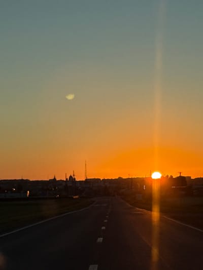 A vertical, high-angle, full shot captures a sunset over the town of Morozovka, Russia. The sky transitions from a light blue at the top to a vibrant orange near the horizon. A faint, circular, textured anomaly appears in the upper left quadrant of the sky. The sun, a bright white orb, is partially obscured by the horizon, casting a strong vertical lens flare down the center of the image. Silhouetted against the sunset is the town's skyline, featuring various buildings, including domes and what appears to be a tower. In the foreground, a dark asphalt road with white dashed lane markers stretches into the distance. The road is framed by dark ground, possibly grass or fields, on either side.