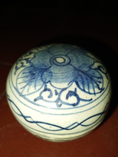 This image, captured in Longmiang, Laos, presents a single, round, lidded ceramic container with blue and white decoration. The object is centered, occupying the lower two-thirds of the frame, and rests on a reddish-brown surface.

**Object:**
The ceramic item features an off-white or cream base glaze with intricate designs rendered in cobalt blue pigment. The domed lid displays a prominent, stylized floral motif, possibly a lotus or peony, as its central element. This flower is depicted with a solid blue wash, a lighter blue outline, and a small, tight spiral at its core. Surrounding the central flower are ornate, flowing scrollwork patterns in fine blue lines. The lower body of the container, visible beneath the lid's edge, is decorated with horizontal bands of continuous, wavy, or intertwined blue lines. The surface of the glaze exhibits a network of fine, irregular crackle lines, and minor dark specks are interspersed across both the white and blue areas.

**Environment:**
The container sits directly on a flat, reddish-brown surface, which appears uniform in texture and color. The upper background is uniformly dark, approaching black, indicating minimal ambient light in that area or a shallow depth of field. The lighting on the object appears to originate from an overhead or front-overhead source, casting no significant shadows within the visible frame and highlighting the texture and curvature of the ceramic piece. There are no people, actions, or interactions depicted.