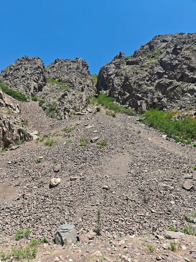 The image depicts a steep, rocky hillside under a clear blue sky. The terrain is covered in loose stones and gravel, with sparse vegetation in patches. Two prominent rock formations rise on either side of a central gully, their surfaces rough and textured. Green bushes and small trees are scattered throughout the scene, particularly along the lower slopes and within the gully. The overall impression is one of a rugged, natural landscape.