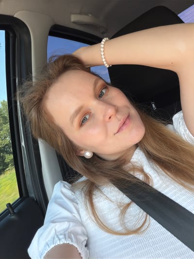 A medium shot captures a woman with long, light brown hair sitting in a car, her right arm raised and hand resting on her head. She is wearing a white, textured shirt and a pearl bracelet. Large pearl stud earrings are visible. Her gaze is directed towards the camera, and her lips are slightly parted. A seatbelt is fastened across her chest. Outside the car window, green foliage and a blue sky are visible, suggesting a daytime setting in a rural or suburban area. The woman's hair is a prominent feature, falling around her shoulders and partially obscuring her right arm. The image appears to be a selfie, taken from a slightly high angle. The location is identified as Tikhonovo, Russia.