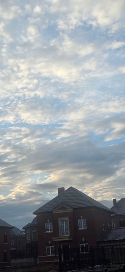 The image shows a view of houses in a suburban area under a cloudy sky. The sky is filled with numerous white and gray clouds, with patches of blue visible. In the foreground, the roofs and upper stories of several houses are visible. The houses are constructed with brick and have dark, pitched roofs. One house in the center is particularly prominent, featuring a red brick facade, white-trimmed windows, and decorative architectural elements. A black metal fence runs along the front of the property. The scene appears to be a residential development, likely in the city of Русская Деревня-2 Кп, Russia, given the context. There are no people visible in the image. The overall atmosphere is overcast and quiet.