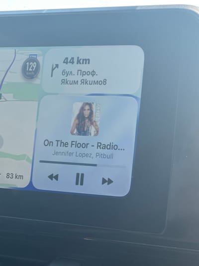 The image shows a car's infotainment screen displaying navigation and music playback information. On the left side of the screen, a map is visible with a speed indicator showing "129 km/h". To the right of the map, navigation details are displayed: "44 km" to "бул. Проф. Яким Якимов" (Prof. Yakim Yakov Street). Below this, a music player interface is shown, featuring the album art of Jennifer Lopez and the song title "On The Floor - Radio..." with artists "Jennifer Lopez, Pitbull". Playback controls, including skip forward, pause, and skip backward buttons, are visible at the bottom of the music player module. The screen is integrated into a car's dashboard, with the surrounding dashboard material being dark gray. The lighting suggests it is daytime. The text "Malenovo, Bulgaria" is not directly visible in the image, but the street name "бул. Проф. Яким Якимов" is a known street in Malenovo, Bulgaria, suggesting the location.