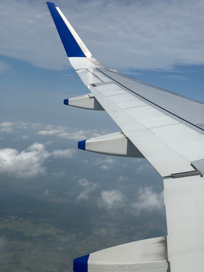 The image captures an aerial view from an airplane window, focusing on the airplane's wing and the landscape below. The wing, predominantly white with blue accents at its tips, extends diagonally across the frame. The sky is a mix of blue and white clouds, some appearing dense and gray, suggesting overcast conditions at lower altitudes. Below the clouds, a patchwork of green fields and a small village or town, likely Mohan, India, is visible. The village appears as a cluster of buildings nestled within the rural landscape, with roads or paths subtly discernible. The overall scene depicts a typical view during an airplane flight over a populated rural area.