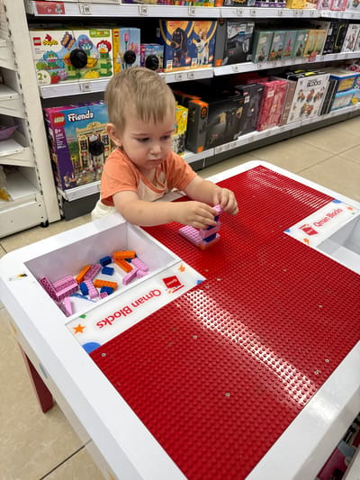 In a retail environment, a young boy is engaged in playing with building blocks. He is positioned at a white and red play table, actively assembling pink and blue blocks. The table has a built-in storage compartment filled with more blocks. The shelves in the background are stocked with various toy boxes, including LEGO Duplo and LEGO Friends sets. The location is identified as the Detsky Mir store in Ramenskoye, Russia.