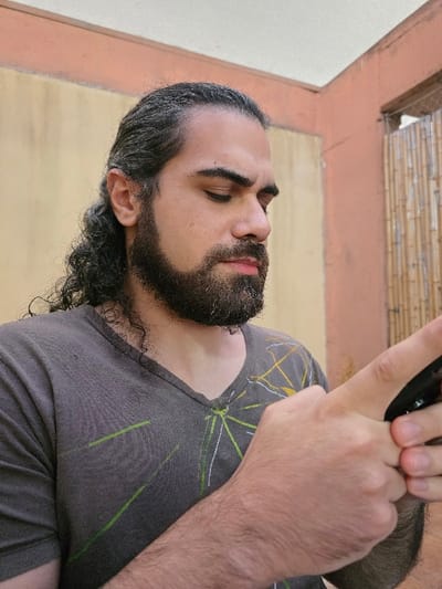 An adult male is depicted from the mid-chest up, located in the city of Villa San Andrés, Argentina. He possesses dark, curly, shoulder-length hair, pulled back, with visible graying, and a full dark beard and mustache, also exhibiting graying. His gaze is directed downwards, suggesting focus on an object. He wears a dark gray V-neck t-shirt adorned with thin, abstract lines in light green, white, and yellow/gold. Both hands are actively engaged in holding a dark, rectangular electronic device, likely a smartphone. The background features a wall with an upper light beige section and a lower orange-brown section. To the far right, a vertical screen or barrier constructed from light brown bamboo or reed-like poles is partially visible.
