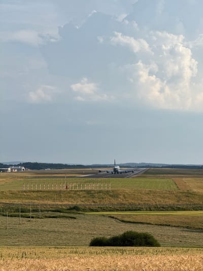An airplane is on a runway at Hahn Airport in Hahn, Germany. The airplane is facing away from the camera. The sky is cloudy. In the foreground, there are fields of crops. There are also some buildings and trees in the distance.