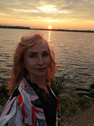 A woman with pink hair is posing for a selfie by a body of water at sunset. The sun is low in the sky, casting a warm orange and yellow glow across the water and clouds. The woman is wearing a colorful patterned jacket over a black top. She is smiling and looking at the camera. The background shows a shoreline with some vegetation and a distant tree line. The overall scene is peaceful and serene.