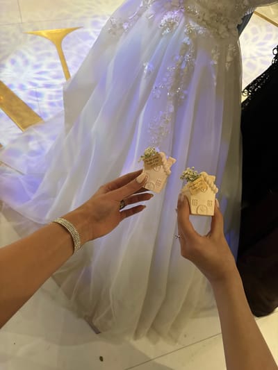 Two female hands hold small, house-shaped favors with tags attached, positioned against the backdrop of a white wedding dress. The favors are beige, and adorned with tiny white flowers and brown labels. The hands are light-skinned, with manicured nails and jewelry; one hand has a ring, and the other a bracelet. The wedding dress is detailed with floral embellishments and is draped over what appears to be a golden support structure. The scene is illuminated with a pattern of geometric shapes, cast on the floor, and suggests an indoor setting. The location of Masis, Armenia is not directly discernible from the image, but the context of a wedding, favors written in Armenian, and the reference to Masis implies a connection.