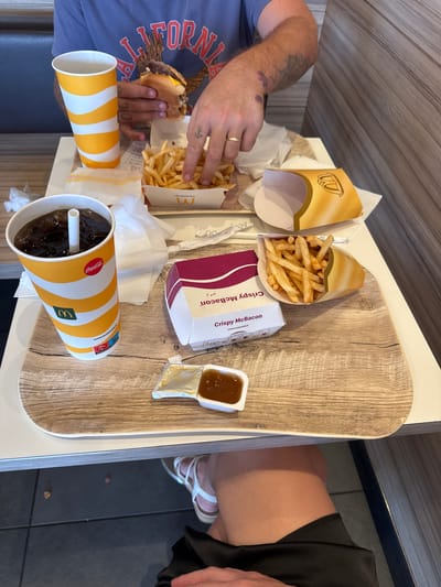 Two people are seen at a McDonald's restaurant in Carini, Italy, sharing a meal. The table is laden with typical McDonald's fare, including a burger, french fries, and two drinks. One person, wearing a blue t-shirt with "California" emblazoned across it, is actively holding the burger and reaching for the fries. The other person, partially visible and wearing a black skirt and white sandals, has their legs extended. The table surface has a wood-grain texture.
Visible text includes "Coca-Cola," "I'm lovin' it," "Crispy McBacon," and a McDonald's logo on the cup and packaging. There's also a web address: "www.mcdonalds.it." The scene appears to be indoors, given the lighting and interior setting of a fast-food establishment. The overall impression is one of a casual meal being enjoyed by two individuals.