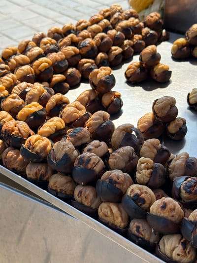 A close-up, high-angle shot shows a large pile of roasted chestnuts for sale on a metal tray, likely in the Fatih district of Istanbul, Turkey, given the typical street food scene. The chestnuts are split open, revealing their soft, golden-brown interiors, with some exhibiting slightly charred edges from the roasting process. The tray is filled with many individual chestnuts, some overlapping, arranged in a casual, abundant manner. The background is slightly blurred, showing a tiled surface and hints of other street elements. The lighting suggests it is daytime, casting subtle shadows on the chestnuts.